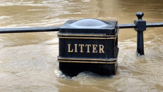 A litter bin surrounded with flood waters, the water rising above the litter sign.