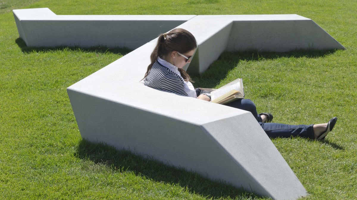 woman reading a book on the grass next to the bench