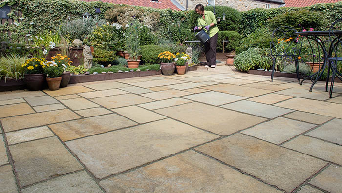 a stone paving patio, with a black metal set of chairs and a table to the right, in the background a woman waters plants