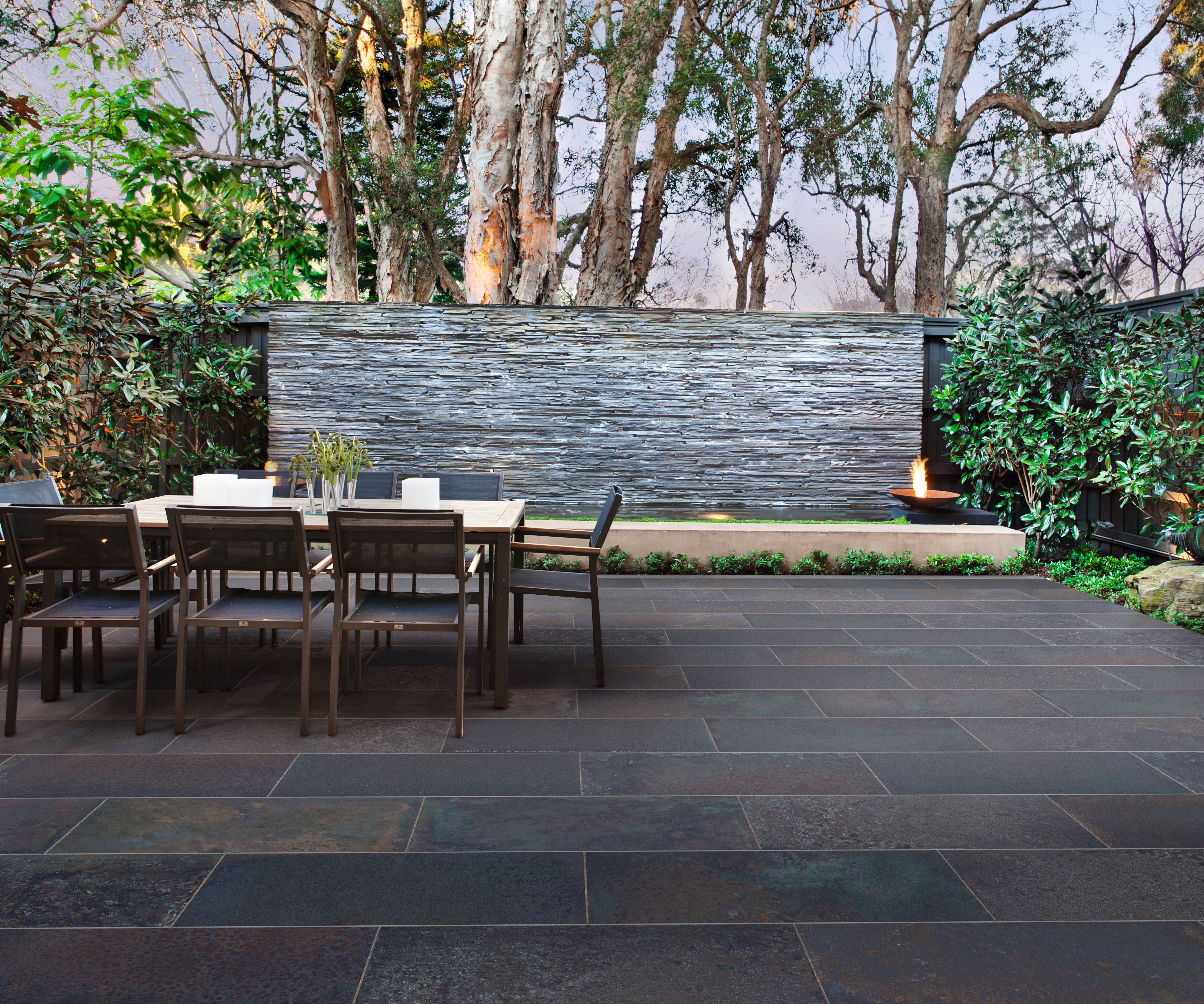 A dark-tiled patio with a table and chairs in front of a water feature